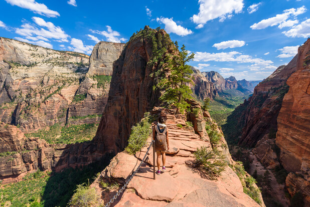 A person hikes along a narrow ridge trail with chains for safety, surrounded by steep cliffs and a vast canyon landscape under a bright, cloudy sky.